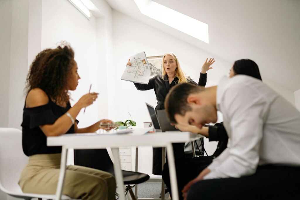 Frustrated manager gesturing with documents as employees show signs of stress and disengagement in a modern office setting.