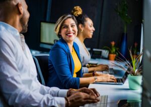 Three diverse professionals smiling and working on laptops in an inclusive office environment.
